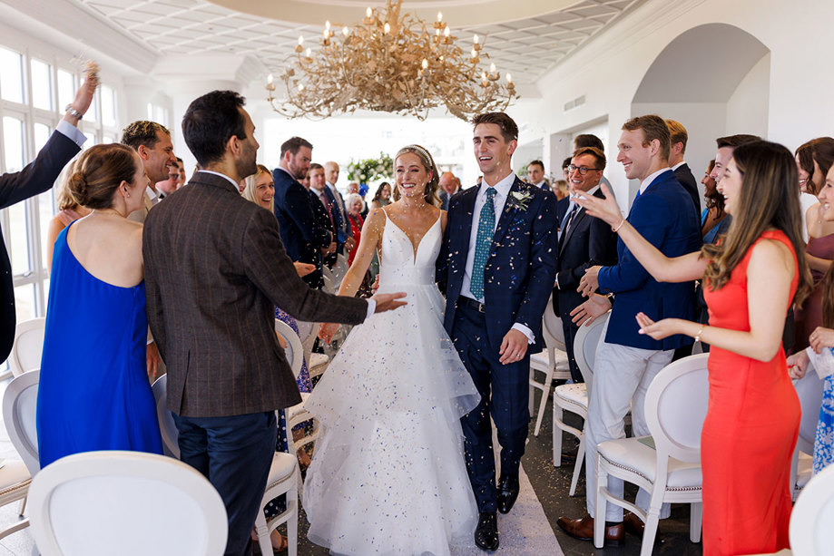 A jubilant bride and groom walk hand in hand, smiling widely as they are showered with flower petals by enthusiastic guests during their Duke Photography captured wedding ceremony at the Old Course Hotel