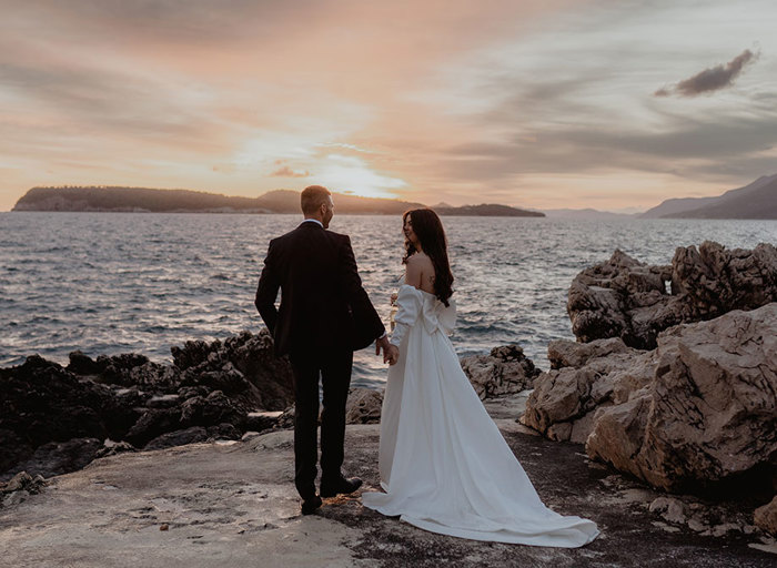 a bride and groom standing on rocks by the sea as the sun sets