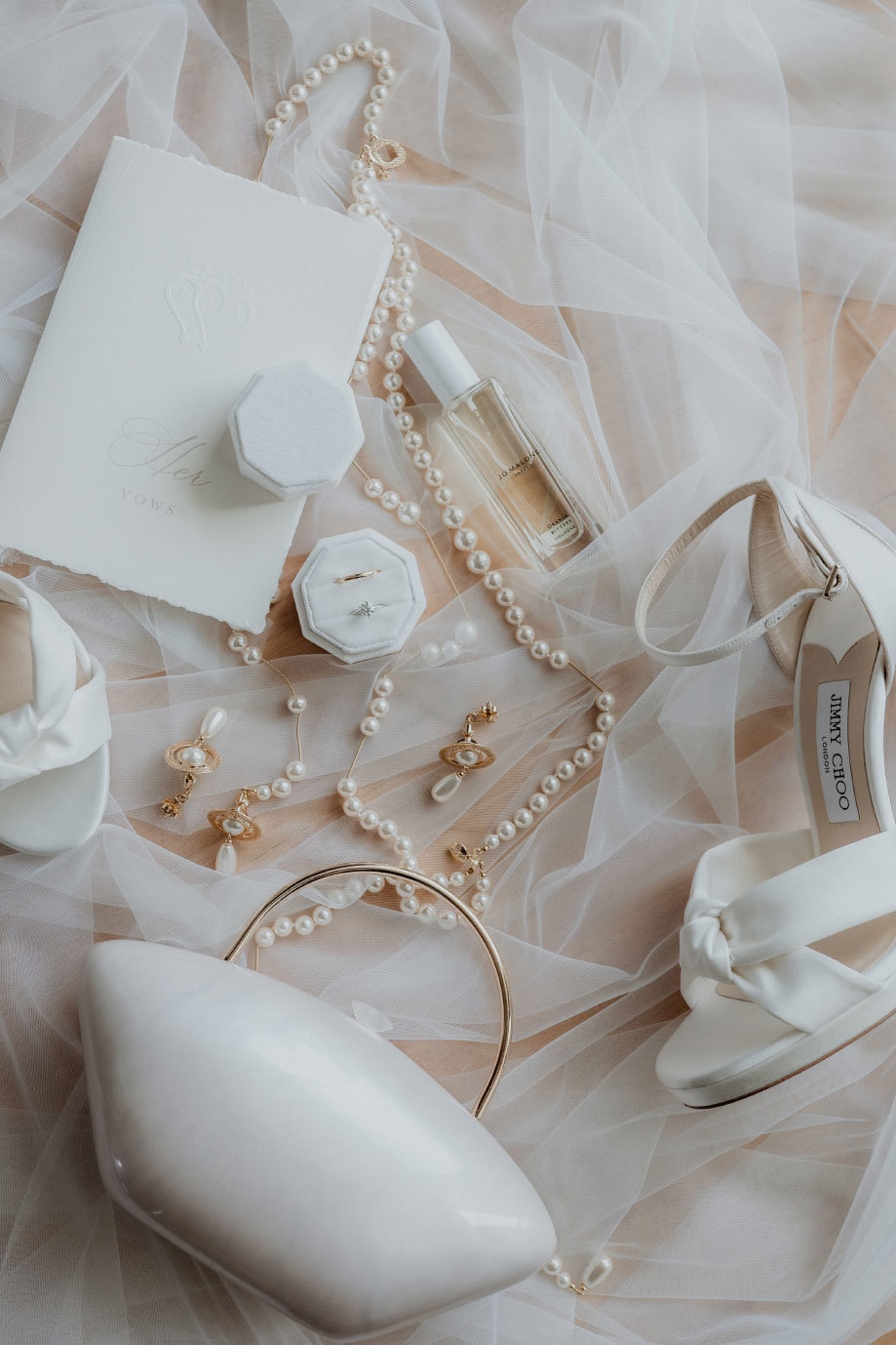 a flat lay of white wedding shoes, hard case bag with gold handle, Vivienne Westwood pearl and gold jewellery, rings and perfume sitting on a white tulle veil