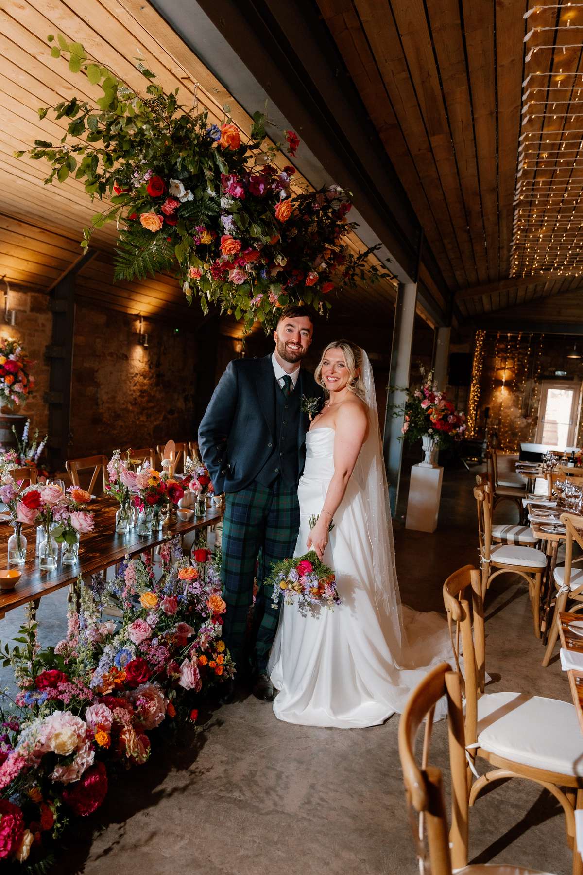Bride and groom portrait beneath a suspended floral installation in the rustic reception barn at Falside Mill
