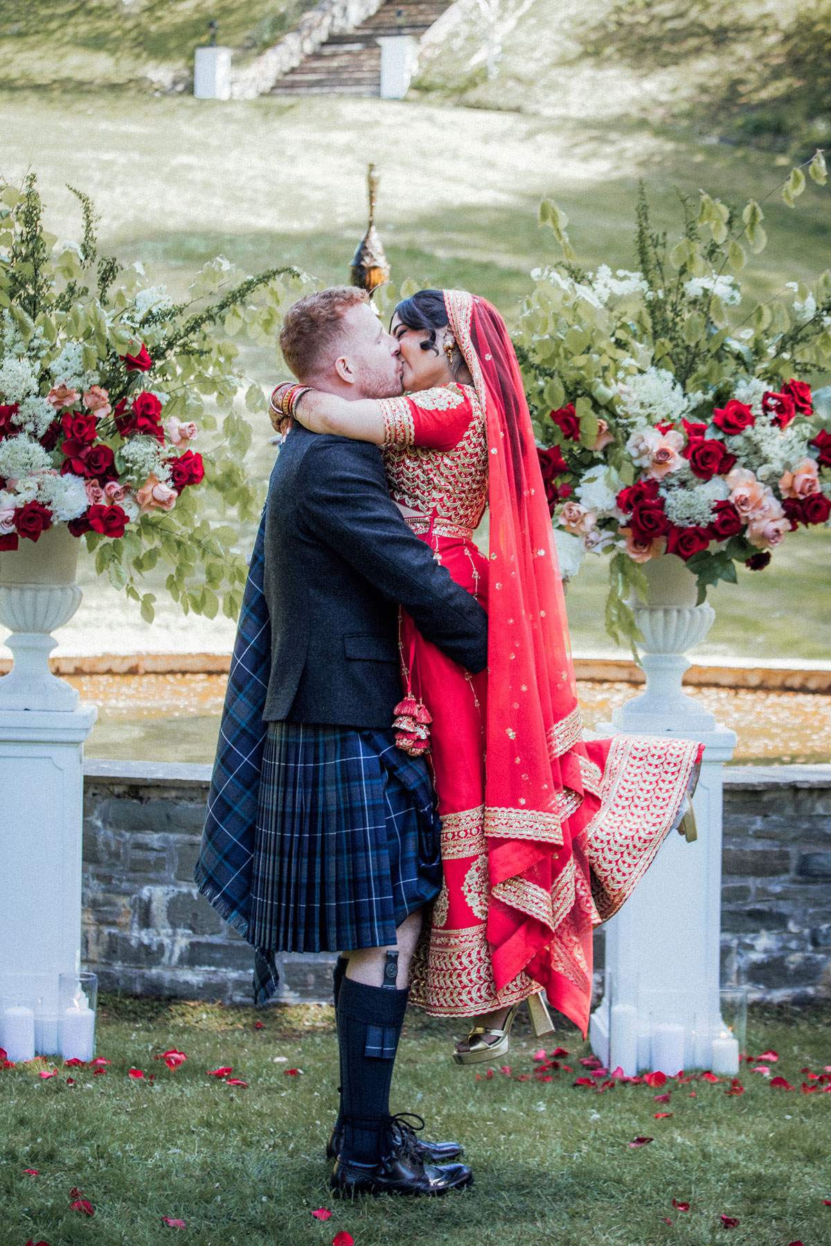 Bride in a red Indian wedding dress and groom in a Scottish kilt sharing a kiss in front of the fountain at Atholl Palace Hotel