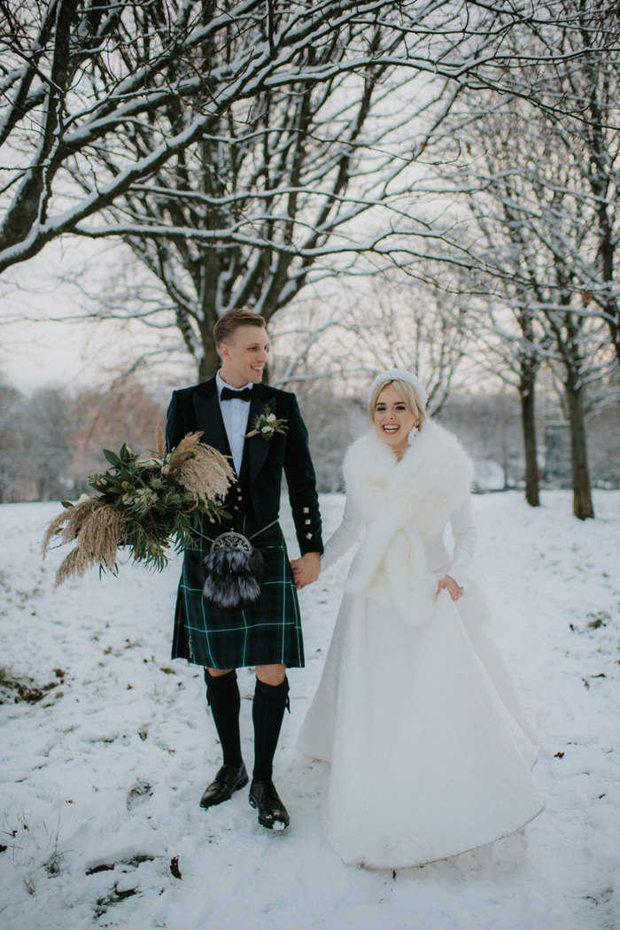 A bride in the snow wearing a white dress and white fur wrap holding hands with a groom wearing a dark kilt