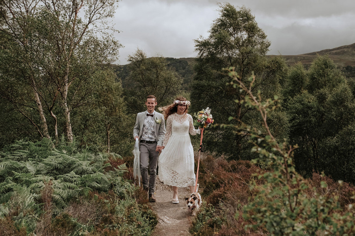 bride and groom walking holding hands with a Jack Russell dog on a lead along a path surrounded by trees