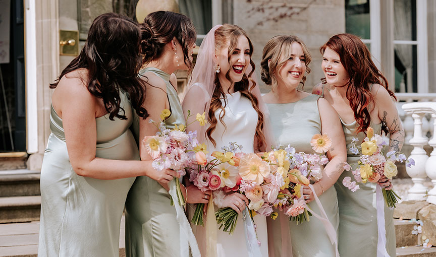Bride and bridesmaids holding pastel bouquets and laughing on the steps outside venue Carlowrie Castle.
