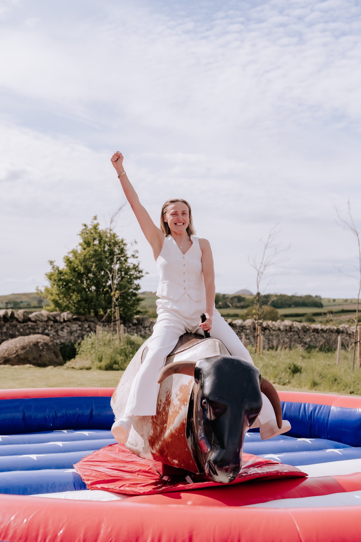 Bride riding a mechanical bull in her white trouser suit at Hannah and Donna’s Harvest Moon wedding.
