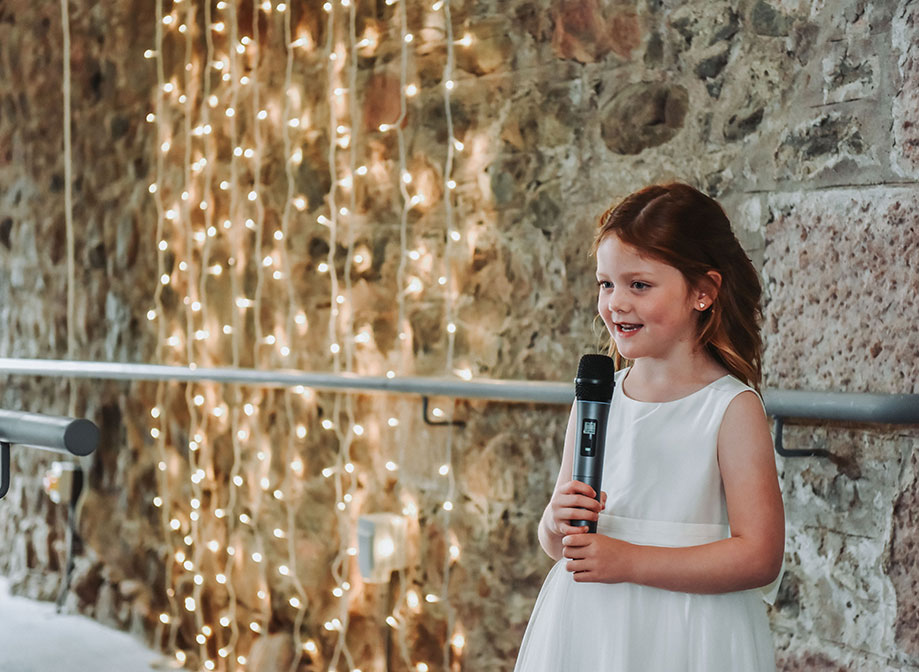 a young girl holding a microphone standing next to a stone wall with strings of fairylights in the background