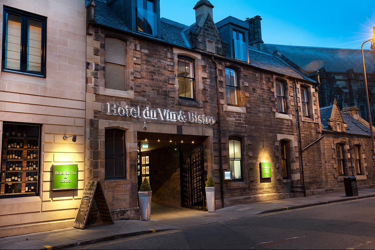 Traditional stone hotel exterior at dusk, with illuminated signage above the entrance and warm lights in the windows