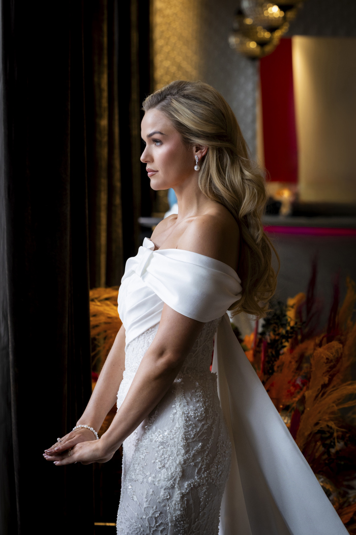 A bride wearing an off-the-shoulder fitted dress turned to the side away from the camera showing her blonde hair in waves down her back