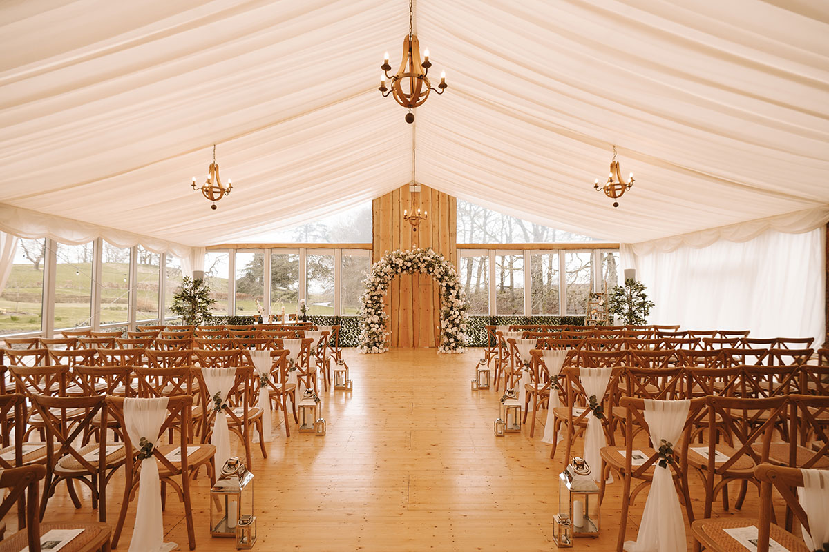 Elegant indoor ceremony setup in a draped marquee with wooden chairs, lanterns lining the aisle, and a floral arch at the altar