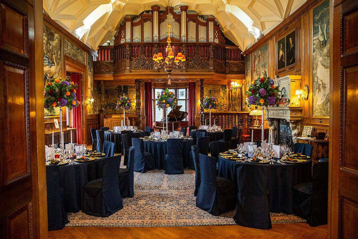 Opulent dining room with high ceilings, tapestries, a grand organ balcony, and round tables dressed in dark linens and floral centrepieces