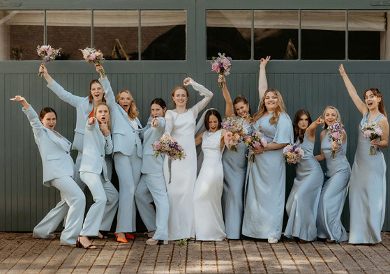 a fun line up of two brides and 10 bridesmaids wearing blue Whistles trouser suits and blue Six Stories posing against a dark teal painted wooden door