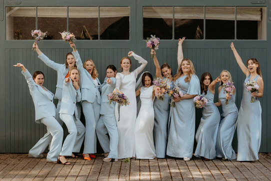 a fun line up of two brides and 10 bridesmaids wearing blue Whistles trouser suits and blue Six Stories posing against a dark teal painted wooden door
