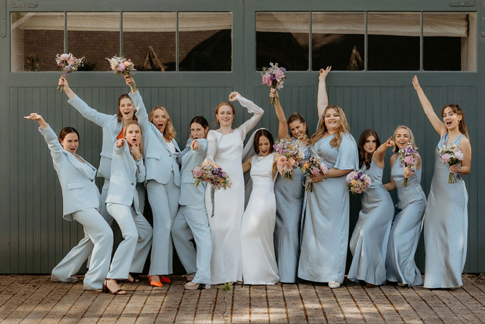 a fun line up of two brides and 10 bridesmaids wearing blue Whistles trouser suits and blue Six Stories posing against a dark teal painted wooden door