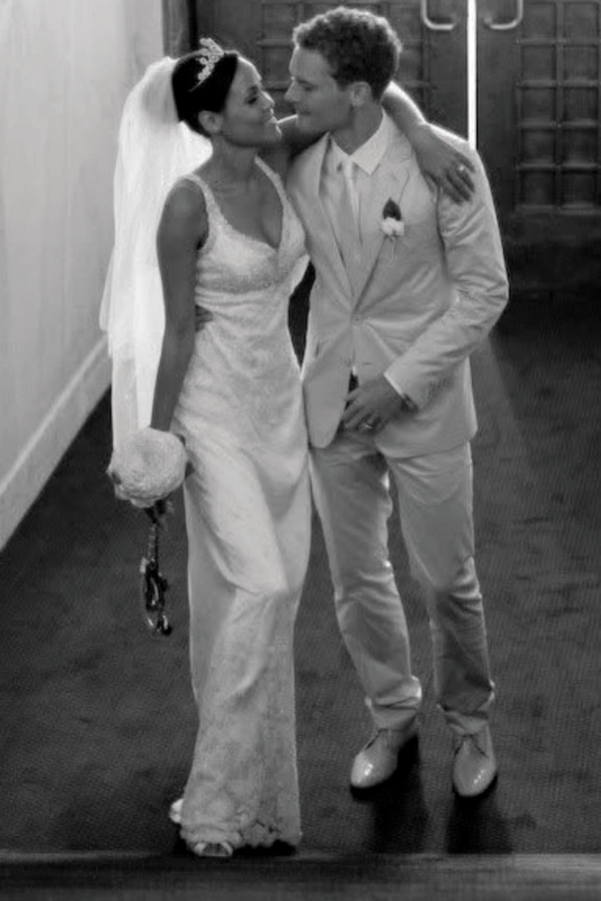 Black and white image of bride and groom walking arm in arm indoors on their wedding day; the bride wears a lace gown with veil and tiara, and the groom wears a light-coloured suit