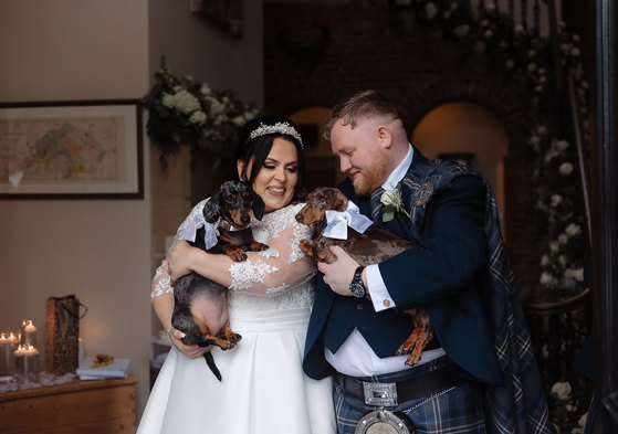 A joyful bride and groom in wedding attire, holding two dachshunds dressed with bows, share a laugh in a warmly lit Logie Country House with floral decorations