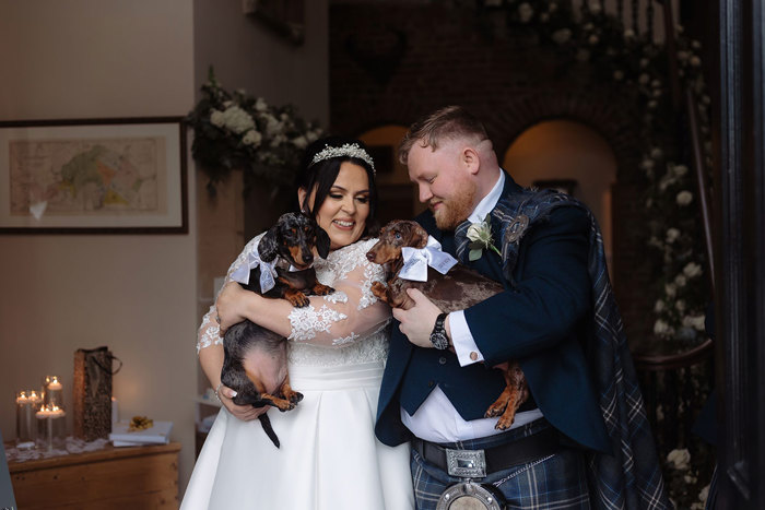 A joyful bride and groom in wedding attire, holding two dachshunds dressed with bows, share a laugh in a warmly lit Logie Country House with floral decorations