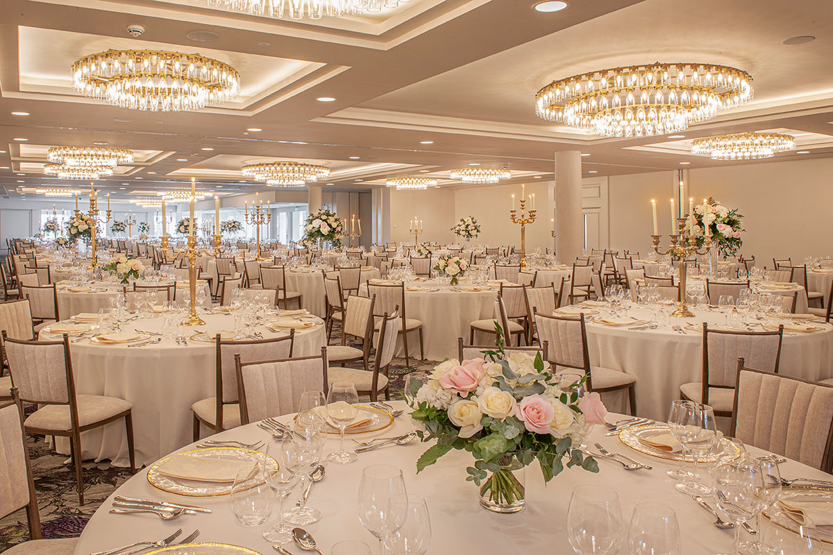 Large wedding reception room set for dinner with round tables, floral centrepieces and modern chandeliers