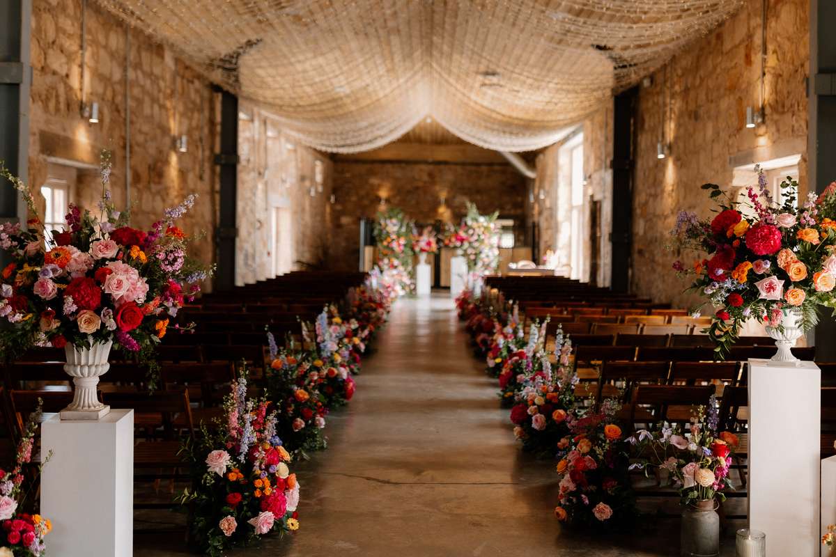 Wedding ceremony setup at Falside Mill with colourful floral aisle arrangements, stone walls and fairy lights overhead