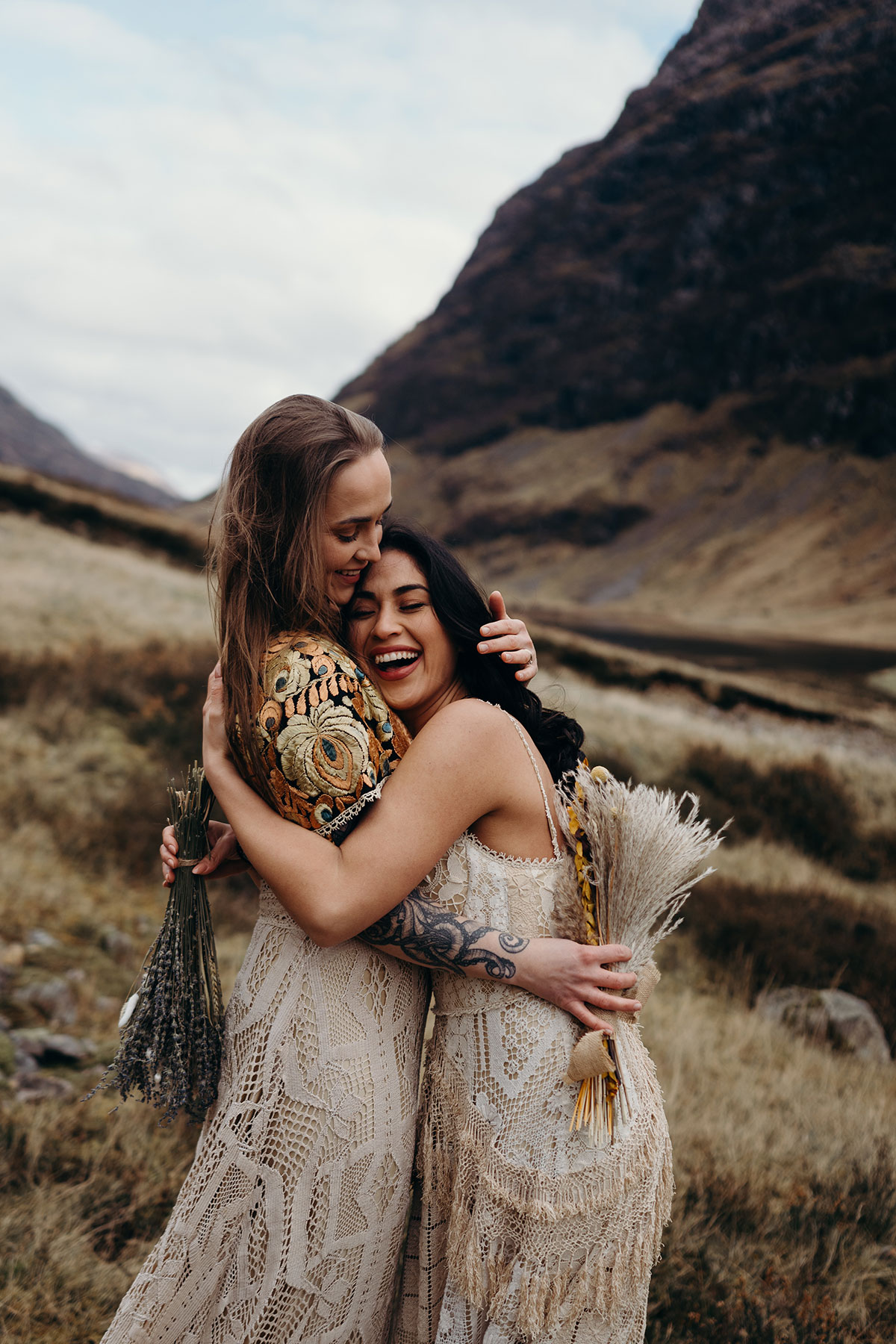 Couple embracing and laughing during a boho Glencoe elopement shoot, holding dried flower bouquets in the Scottish Highlands.