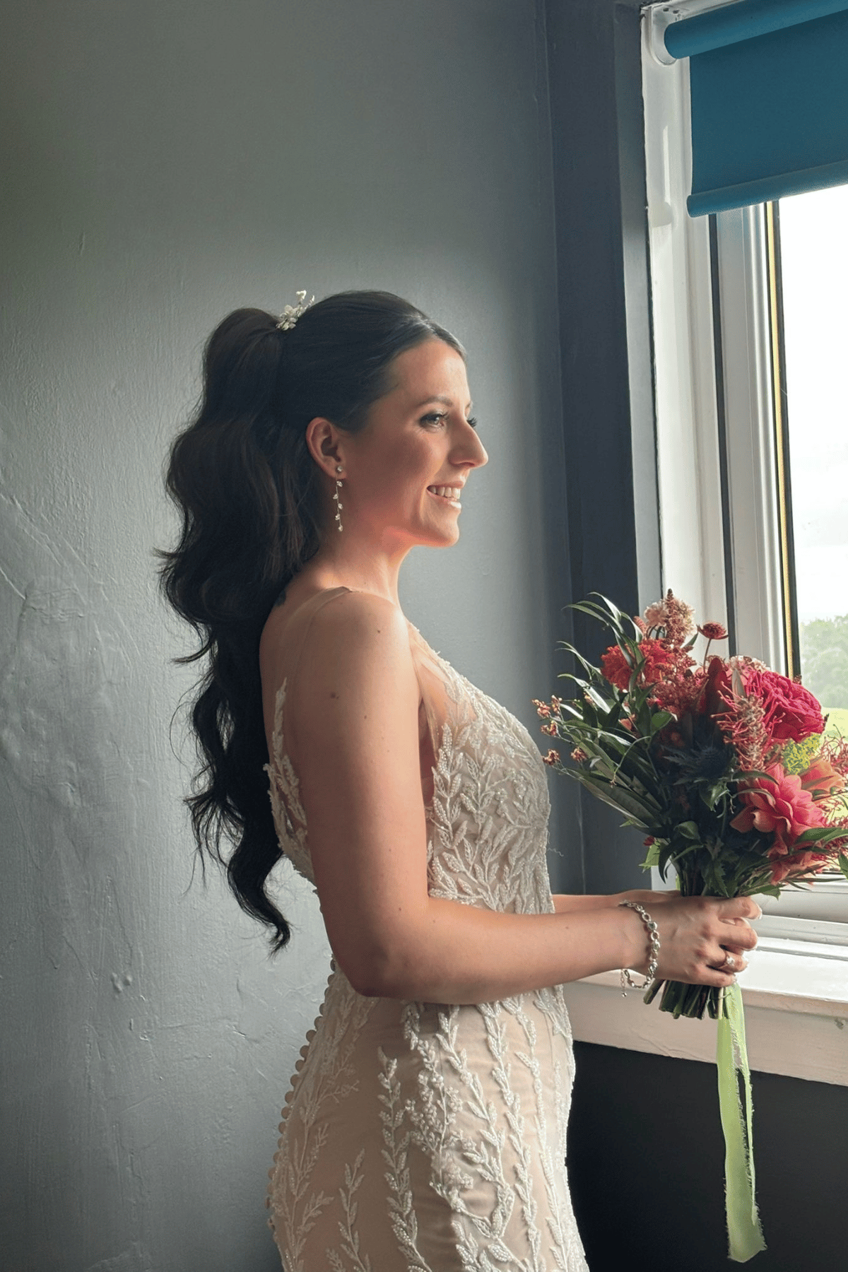 Bride with a long wavy ponytail wedding hairstyle holding a colourful bouquet by a window