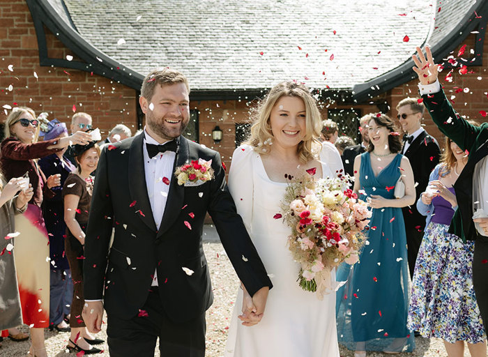 an elated bride and groom walking hand in hand through two rows of wedding guests while being showered in red and white confetti