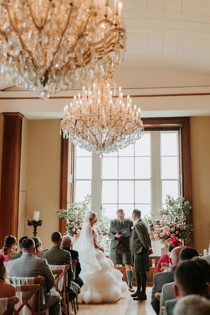 audience view of bride of wedding ceremony with bride in a large ballgown and groom plus celebrant in kilt outfits all three stood at the top of the aisle