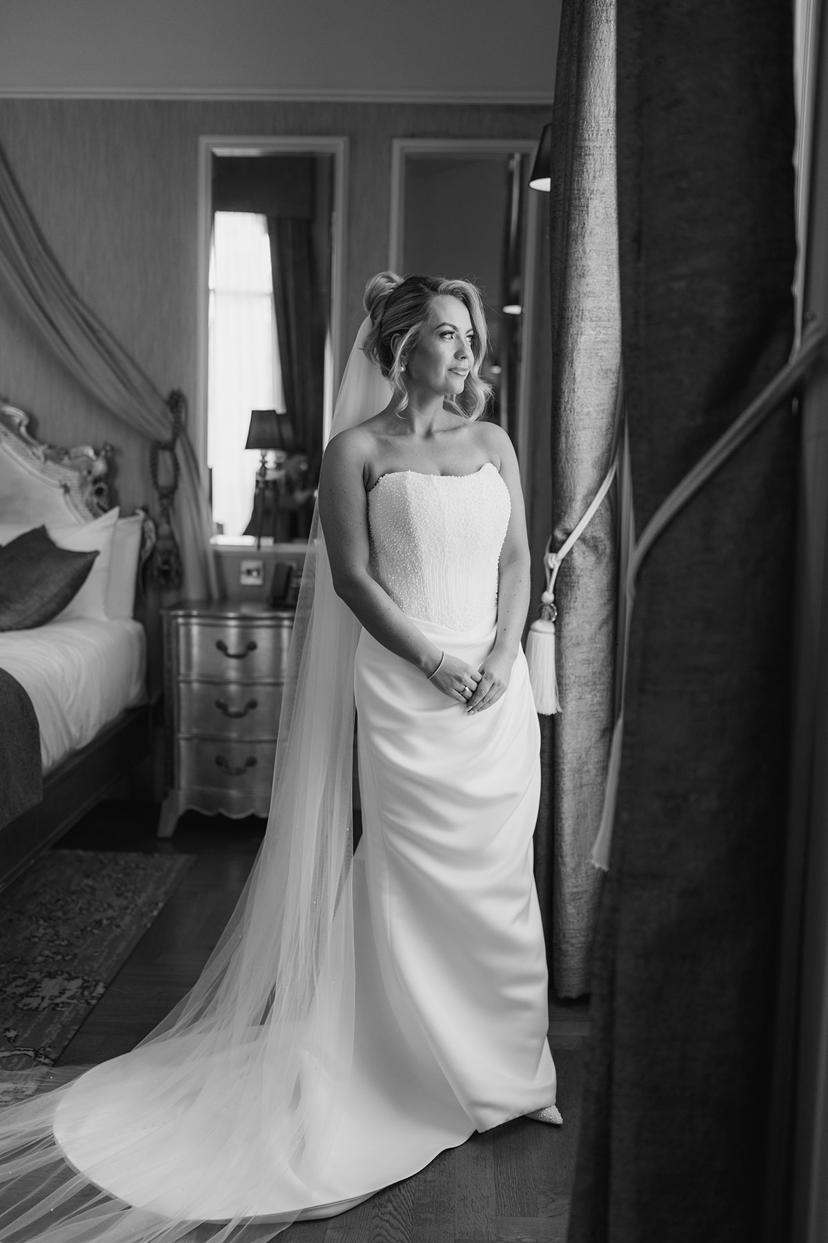 Black and white portrait of the bride in her wedding dress and veil standing beside the window in her bridal suite