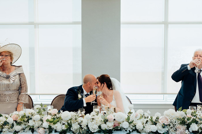a bride and groom kiss and clink champagne glasses while sitting behind a row of white and pale pink flowers 