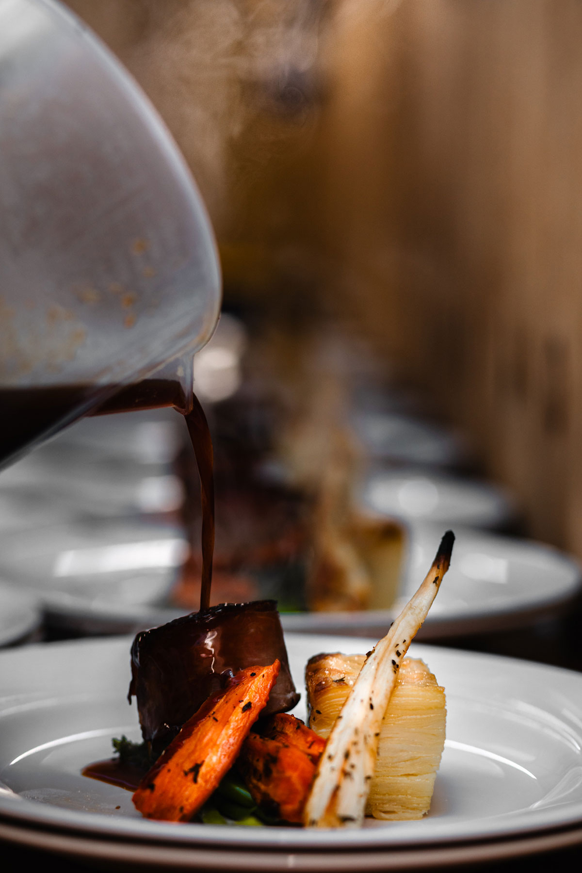A close-up of rich gravy being poured over a plated beef dish with roasted carrots and layered potatoes at a wedding meal