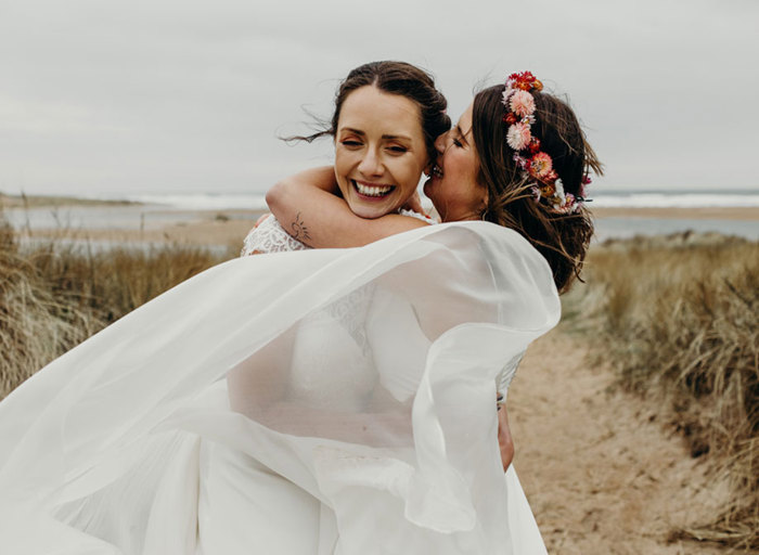 a bride wearing a flower crown kisses another bride on the cheek. They are standing on a sandy path surrounded by scrubby tall grass with the sea in the background