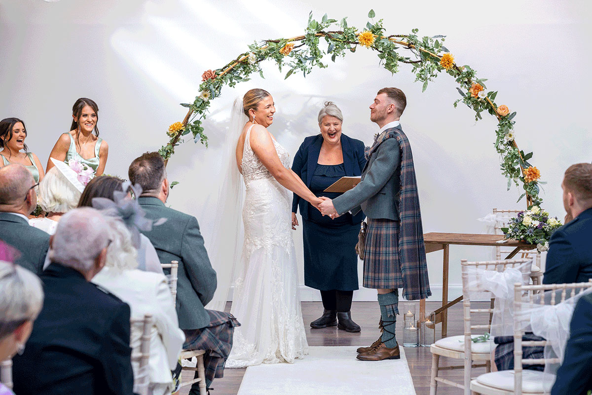 Scottish wedding ceremony at Dr Bell’s Baths led by celebrant Yvonne from Fuze Ceremonies, with the couple smiling under a floral arch surrounded by guests.