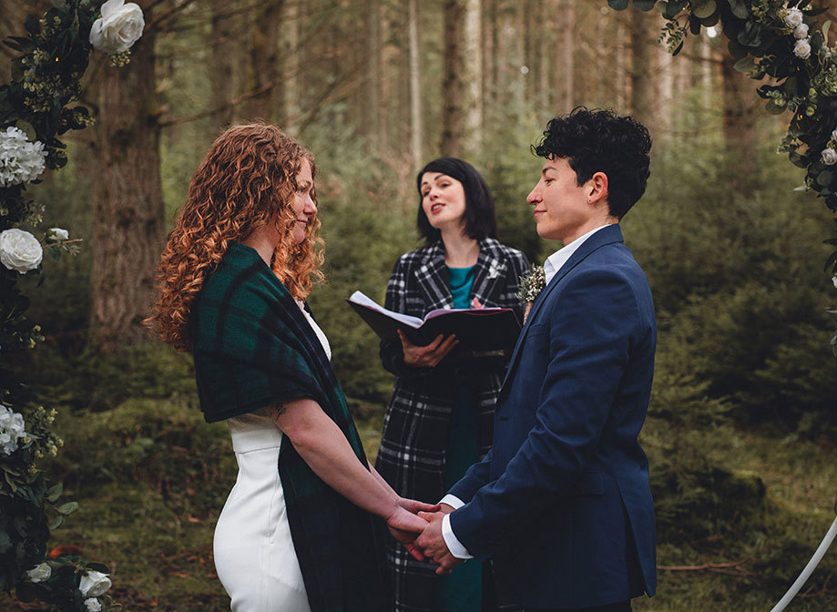 Wedding ceremony in a forest between two women with a female celebrant holding a book 