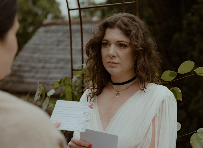 an emotional looking bride looks at another bride who is reading off a small note card