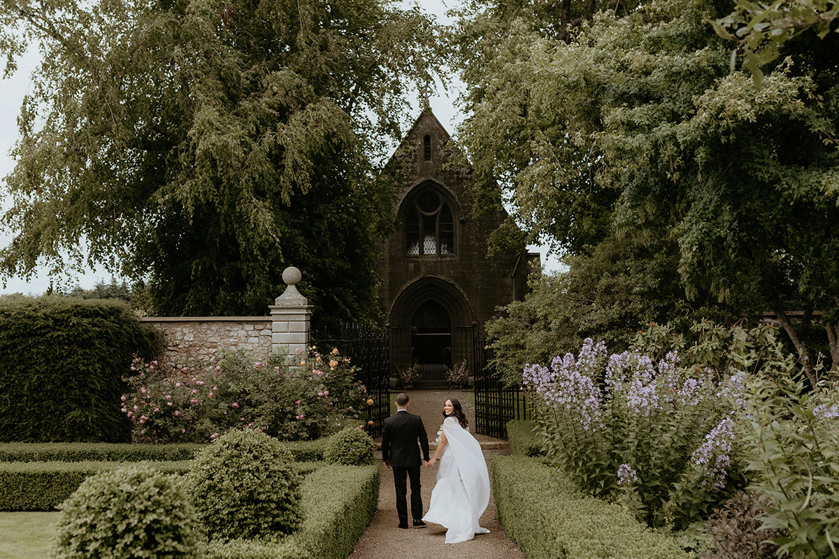 Newlyweds walking through formal gardens towards chapel at Rosebery House and Steading, Midlothian countryside wedding venue.