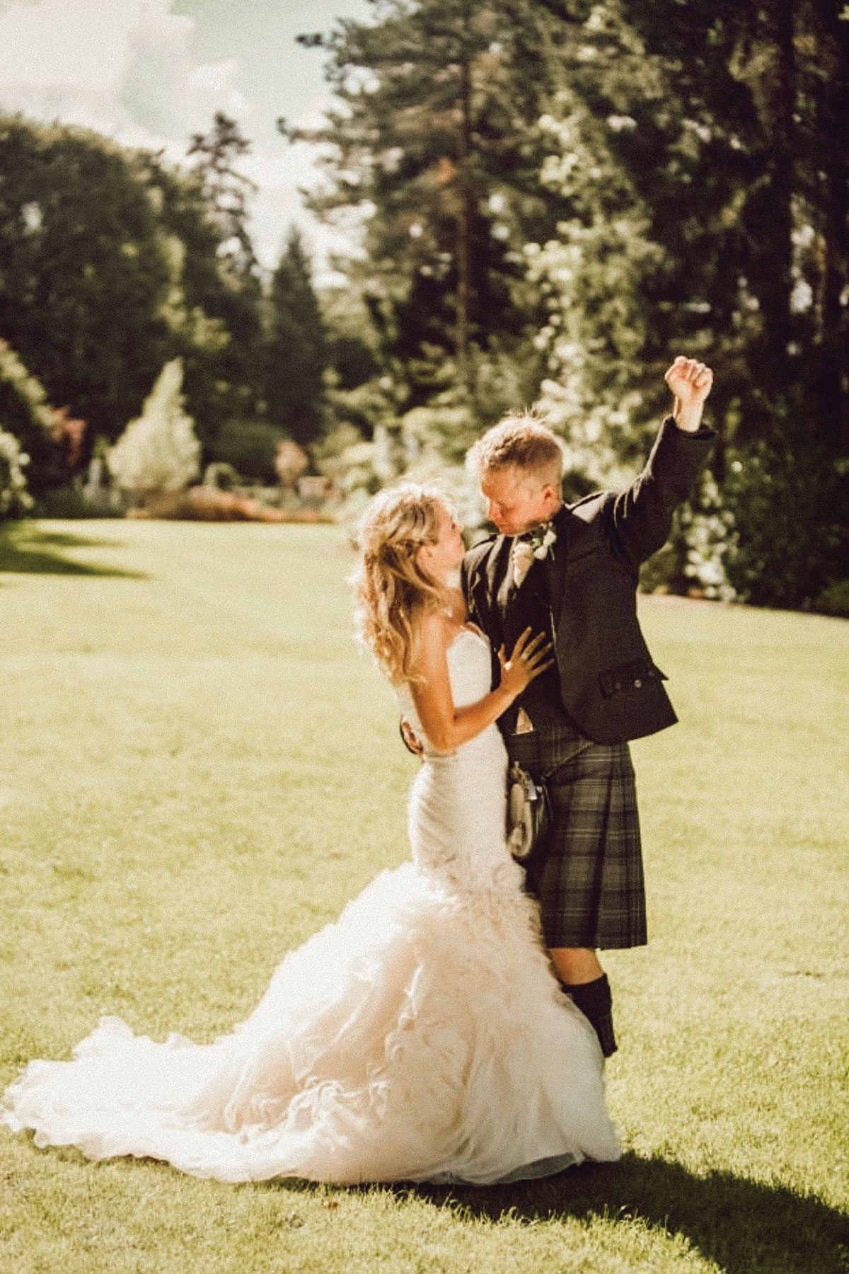Bride and groom embracing on a lawn in warm sunlight; the bride wears a fitted gown with a feathered hem and the groom wears a dark jacket with a kilt