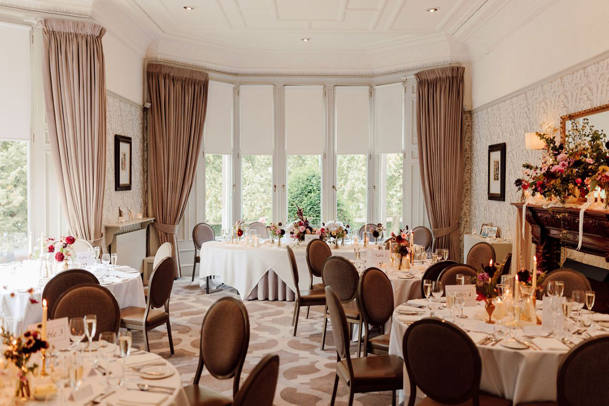 A bright reception room with bay windows, round tables, neutral drapes and floral centrepieces.