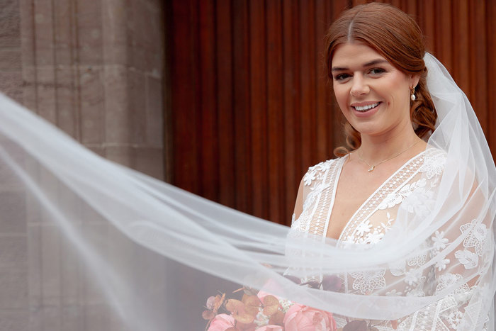 brunette bride smiles as long tulle veil blows out from behind her and partially covers the front of her gown