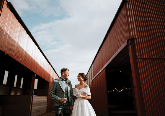 bride and groom stand between two red barns each holding a drink and smiling at each other