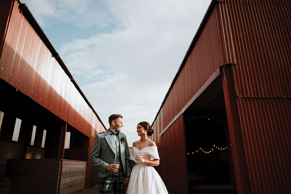 bride and groom stand between two red barns each holding a drink and smiling at each other