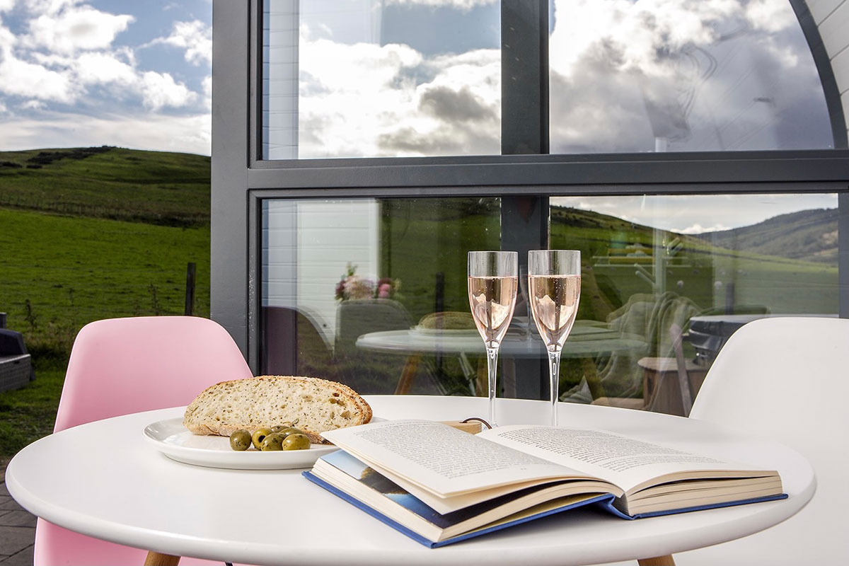 a round outdoor table has an open book, two glasses of fizz and a plate with slices of bread and olives on it – blue skies and green grass can be seen in the distance behind
