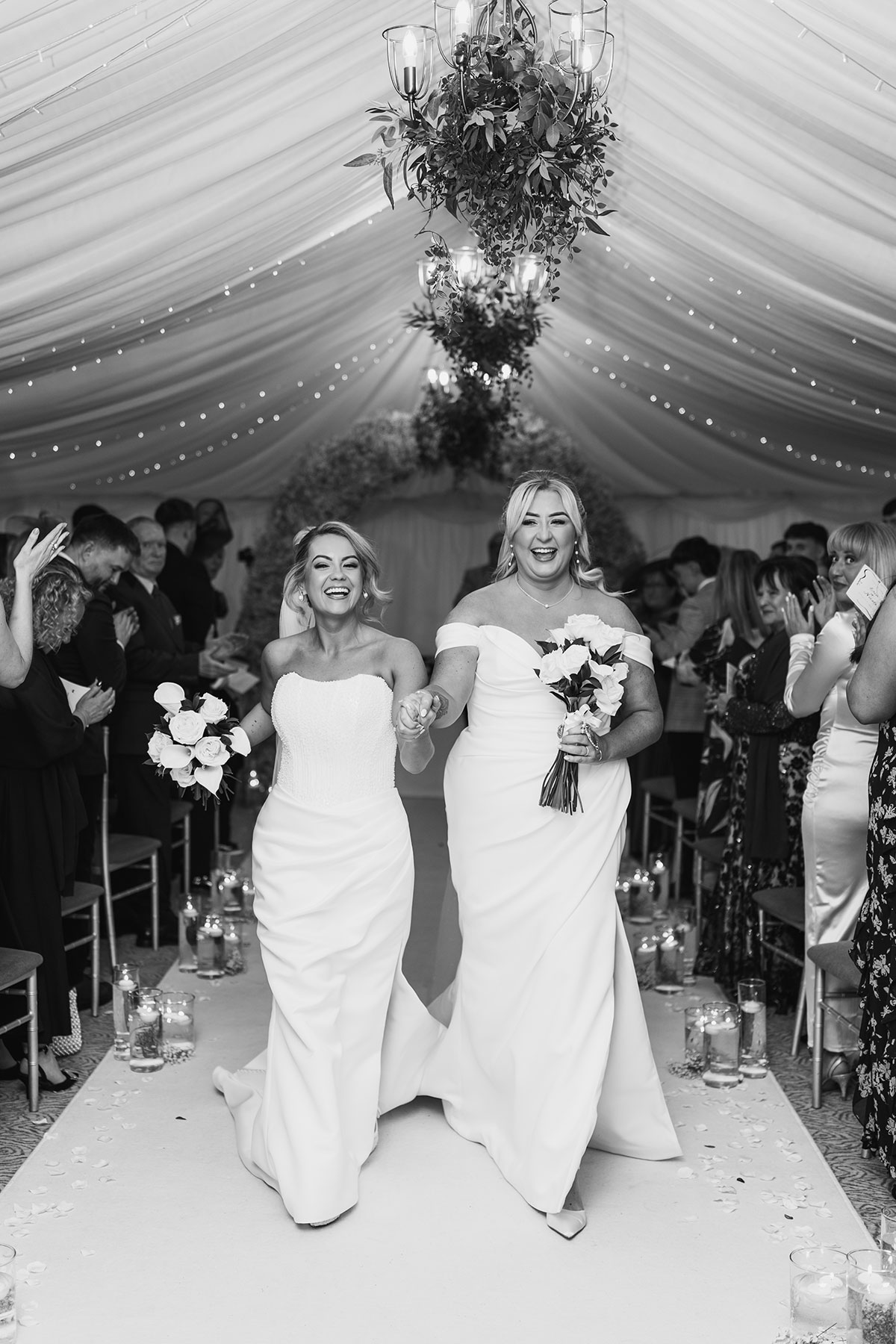 Black and white photograph of two brides walking hand in hand down the aisle after their wedding ceremony as guests applaud