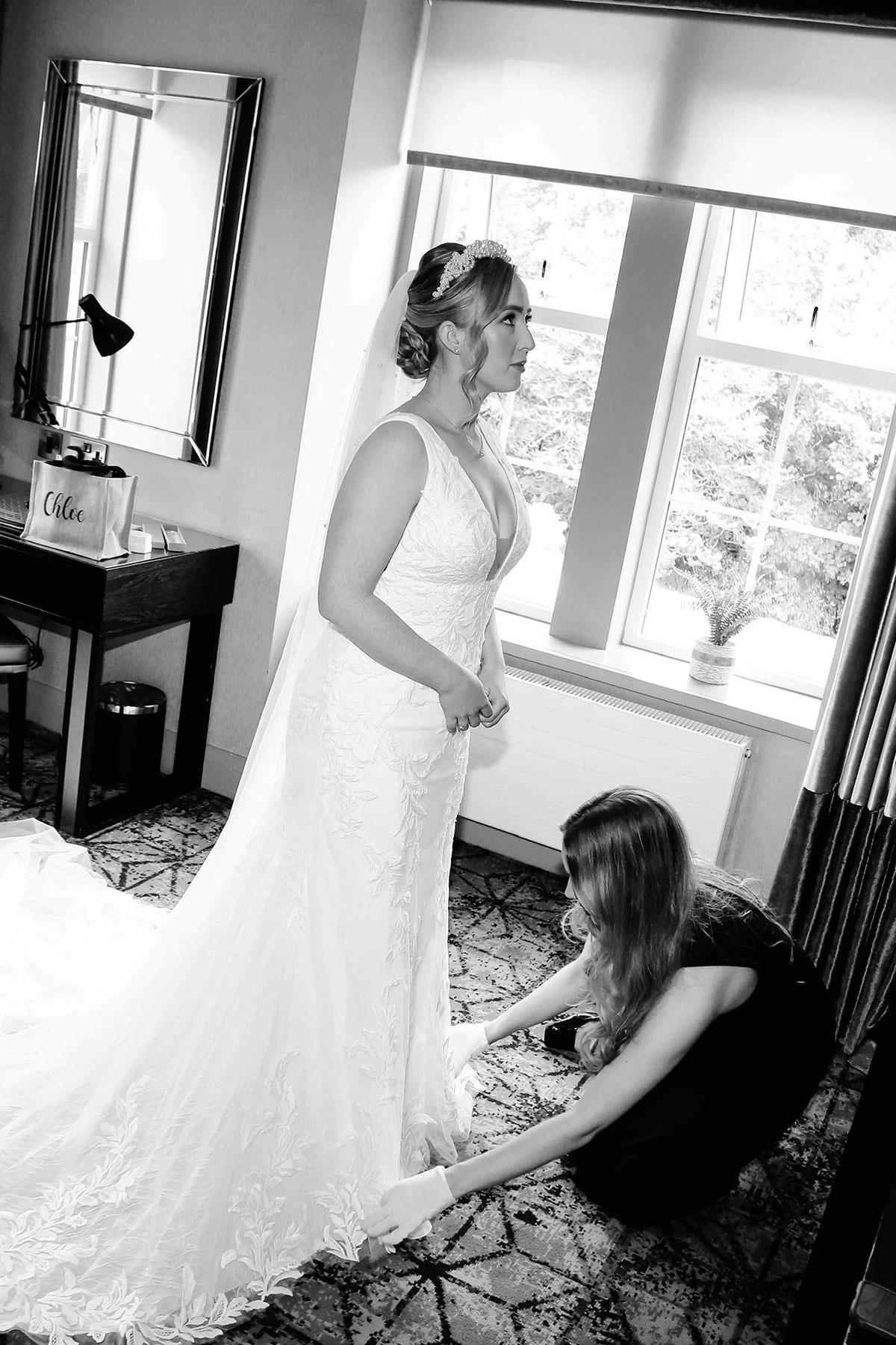 black and white image of a woman in all black outfit, crouched down on carpet, adjusting the hem of a wedding dress with her white gloved hands
