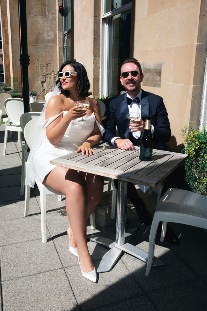 A bride in a short dress sits next to a groom in a navy tuxedo at a table outside, both holding coupes of champagne