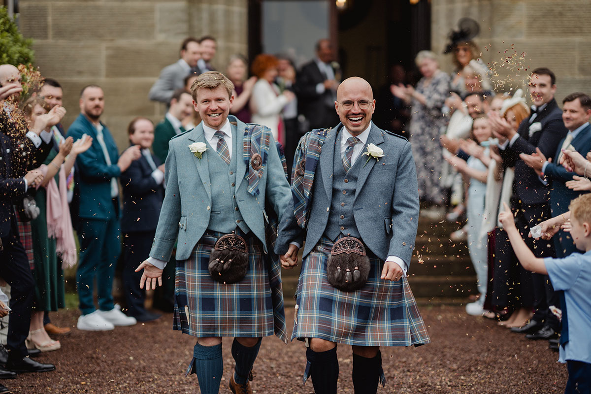 Two grooms in matching blue tweed jackets and tartan kilts walk hand in hand through a confetti shower as guests cheer outside a grand stone building