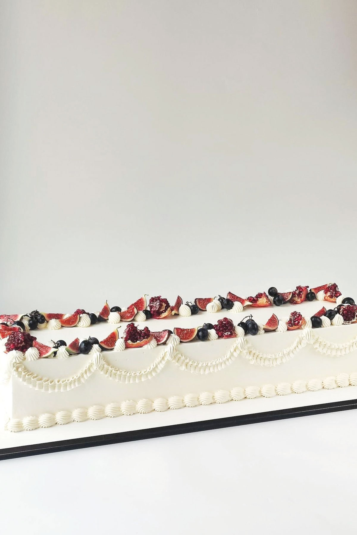 Long rectangular wedding cake decorated with fresh figs, blueberries, pomegranate and piped white buttercream