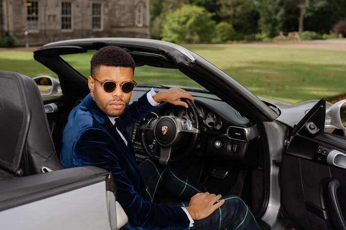 A man wearing sunglasses, a blue velvet jacket and green tartan trousers sits in a silver porche looking back at the camera over his shoulder