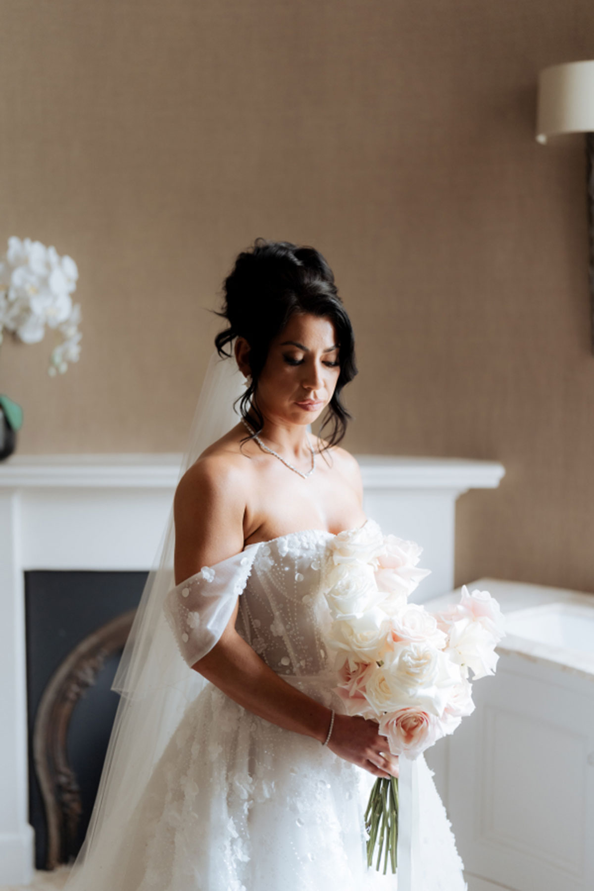 Bride in off-the-shoulder wedding dress holding soft blush and ivory rose bridal bouquet indoors