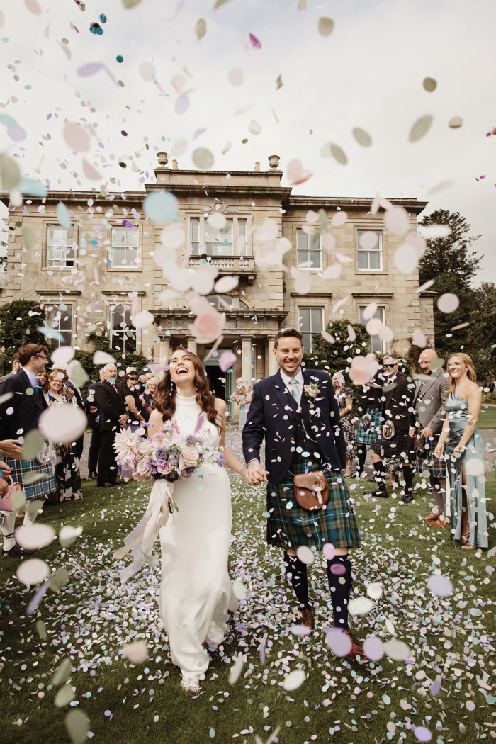 bride and groom hold hands as guests shower them in pastel-coloured confetti during wedding at netherdale house