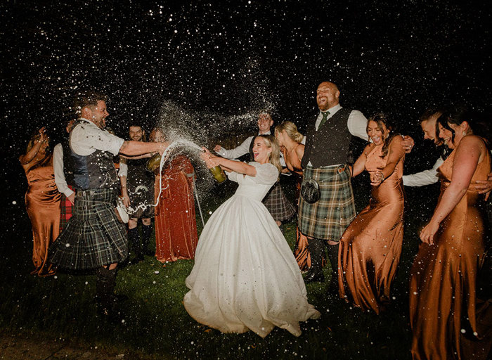 a bride and groom doing a champagne spray at each other in the dark with men in kilts and women in rust coloured bridesmaid dresses cheer in the background 