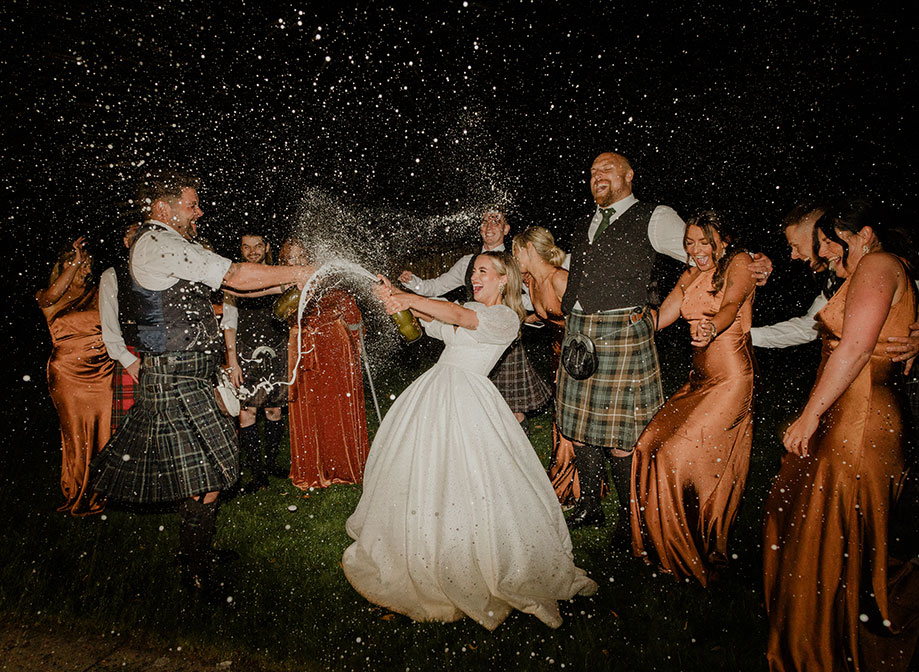 a bride and groom doing a champagne spray at each other in the dark with men in kilts and women in rust coloured bridesmaid dresses cheer in the background